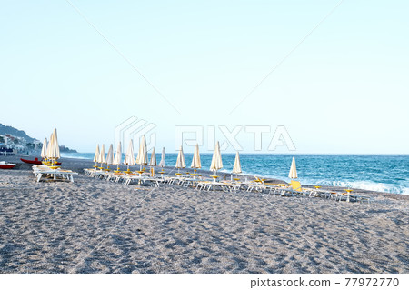 Deserted pebble beach with deck chairs and closed umbrellas in the evening. Sicily as a tourist attraction. Season on mediterranean sea. Ionian sea Deserted pebble beach with deck chairs and closed umbrellas in the evening. Sicily as a tourist attraction. Season on mediterranean sea. Ionian sea 77972770