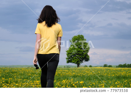 A girl in a yellow T-shirt moves across the field with flowers. Yellow flowers, green grass, blue sky, oak. Travel outside the city. Leisure 77973538