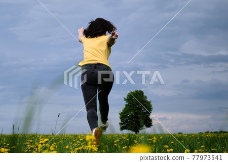 A girl in a yellow T-shirt moves across the field with flowers. Yellow flowers, green grass, blue sky, oak. Travel outside the city. Leisure A girl in a yellow T-shirt moves across the field with flowers. Yellow flowers, green grass, blue sky, oak. Travel outside the city. Leisure 77973541
