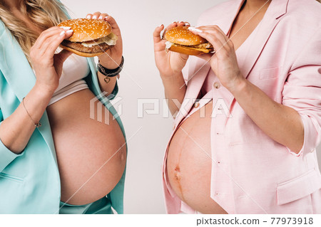 Two pregnant women in suits with hamburgers in their hands on a gray background Two pregnant women in suits with hamburgers in their hands on a gray background 77973918
