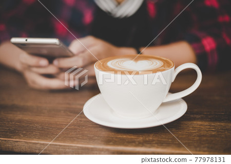 Hipster woman's hand in cafe using mobile phone and texting in social networks with a cup of late art coffee reflection from window outdoors. 77978131
