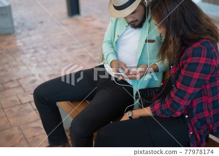 Hipster couple listening music together while sitting on bench and enjoying in urban. 77978174