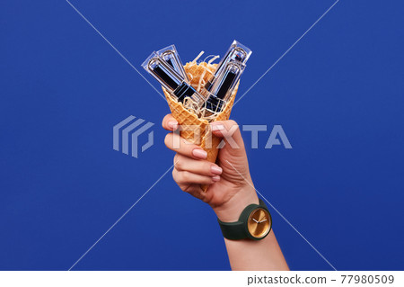 Hand of a female in watch is holding a wafer cone filled with four lipsticks against blue studio background. Cosmetic, makeup. Close up, copy space 77980509