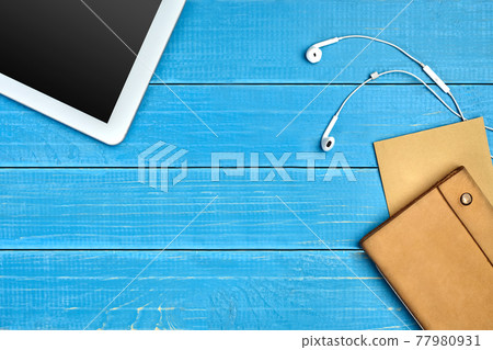 White tablet with black screen, earphones, brown craft paper sheet and leather folder on blue wooden desktop. Close up, copy space White tablet with black screen, earphones, brown craft paper sheet and leather folder on blue wooden desktop. Close up, copy space 77980931