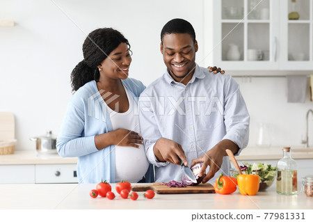 Happy african american pregnant couple cooking together, kitchen interior Happy african american pregnant couple cooking together, kitchen interior 77981331