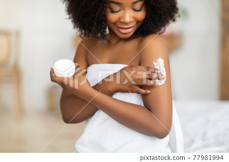 Beautiful black lady wearing towel after bath, applying cream, rubbing in nourishing body butter at home Beautiful black lady wearing towel after bath, applying cream, rubbing in nourishing body butter at home 77981994