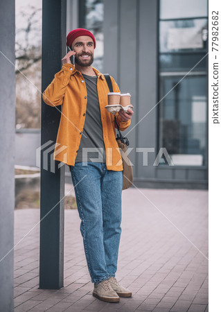 Young bearded man in a red hat talking on the phone and carrying the coffee cups 77982682