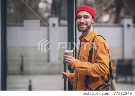 Man in a red hat and orange jacket with a coffee cup in hands looking cheerful 77982849