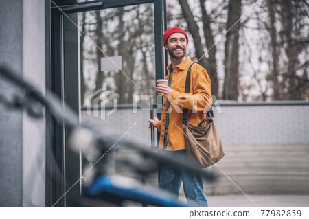 Man in a red hat and orange jacket with a coffee cup in hands looking cheerful 77982859