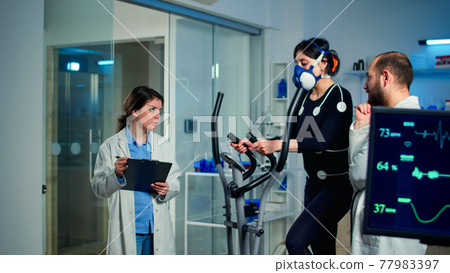Sportswoman running with mask on cross trainer with electrodes attached to body 77983397