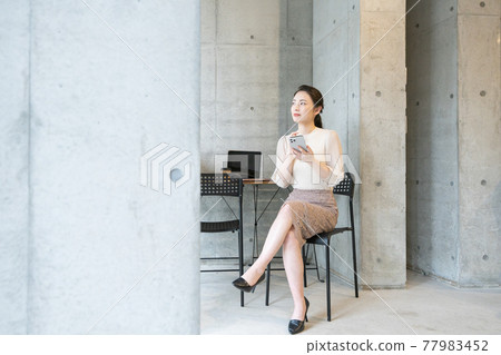 A young woman sitting in a chair in a fashionable office and looking at her smartphone with her legs crossed 77983452