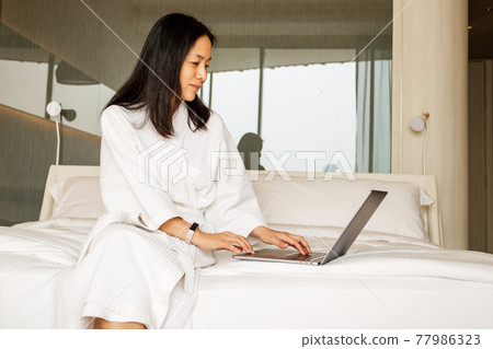 Woman sitting on the bed in dressing gown working laptop. 77986323