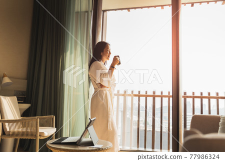 Happy businesswoman in dressing gown stands near the window holding cup of coffee. 77986324