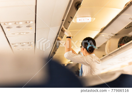 Flight attendant in protective masks and glove closing overhead compartment. Flight attendant in protective masks and glove closing overhead compartment. 77986516