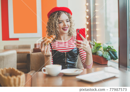 French woman eating croissant and using smartphone in cafe 77987573