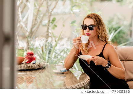 A young woman in an elegant black dress and sunglasses drinks coffee at a table in a cafe A young woman in an elegant black dress and sunglasses drinks coffee at a table in a cafe 77988326