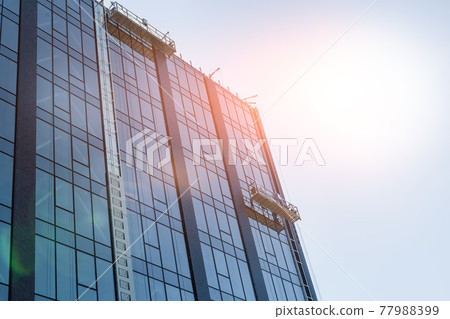 Suspended construction craddle near wall of hightower residentaial building with glass metal modern facade construction site. Engineering urban city development background. Sun shining flare on sky 77988399