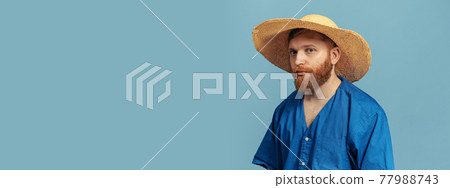 Young red headed and bearded man in straw hat standing isolated over blue bacground. Flyer 77988743