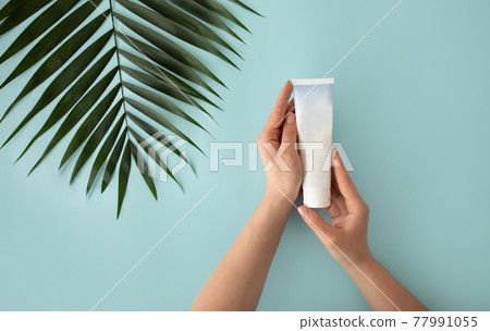Female hands holding white tube for cream, moisturizer, lotion or facial cleanser on blue background with tropical leaf 77991055