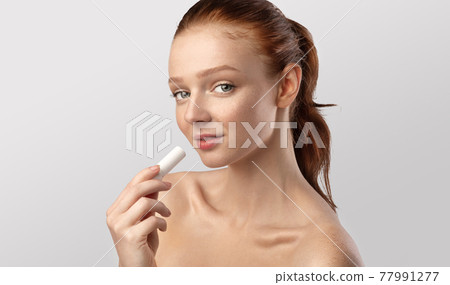 Red-Haired Teenager Girl Using Lip Balm Over Gray Background Red-Haired Teenager Girl Using Lip Balm Over Gray Background 77991277