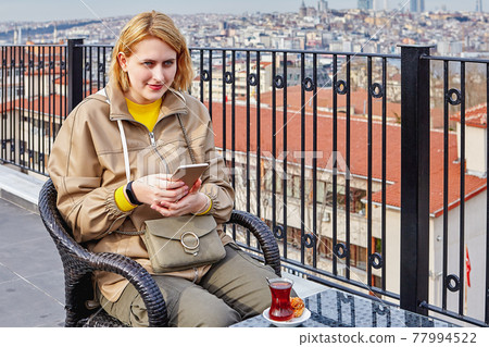 Tea drinking against backdrop of cityscape of Istanbul and online communication with friends using smartphone, young woman is texting on Internet. Tea drinking against backdrop of cityscape of Istanbul and online communication with friends using smartphone, young woman is texting on Internet. 77994522