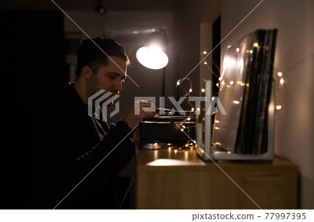 Young unshaven handsome hipster guy puts tonearm on vinyl record on turntable 77997395