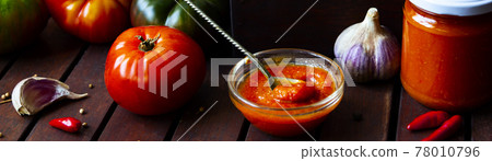 Homemade hot tomato sauce adjika in jars - Tomatoes, chilli pepper, garlic, herbs on wooden table. Homemade hot tomato sauce adjika in jars - Tomatoes, chilli pepper, garlic, herbs on wooden table. 78010796