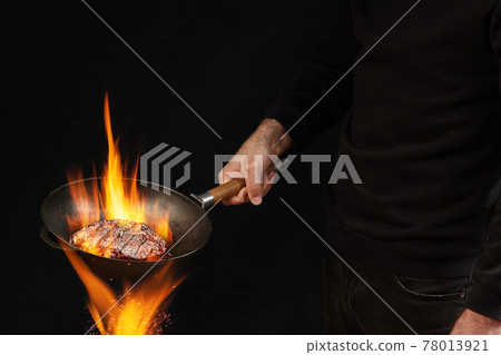 Young fellow dressed in jumper and jeans. He is holding burning wok pan above fire and frying beef steak, against black studio background. Side view Young fellow dressed in jumper and jeans. He is holding burning wok pan above fire and frying beef steak, against black studio background. Side view 78013921