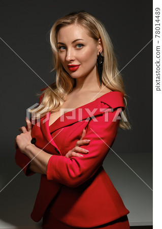 Blonde female, bright make-up and deep neckline, hands folded, in red dress and black earrings. Smiling, posing sideways on gray background. Close up 78014489