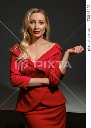 Blonde woman with bright make-up and deep neckline, in red dress and black earrings. She smiling, posing on gray background. Copy space, close up 78014492