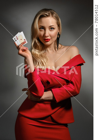 Blonde lady in red dress and black earrings. She smiling, showing two playing cards, posing on gray studio background. Poker, casino. Close-up 78014512