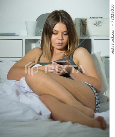 Smiling girl lying and using smartphone on bed in bedroom 78029010