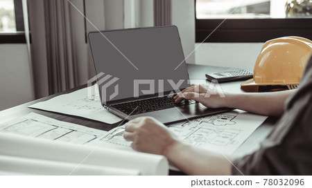 Young man with a laptop plotting a system of building structures in blueprints, Architects or engineers are designing buildings using computers to calculate the physical structure to be correct. Young man with a laptop plotting a system of building structures in blueprints, Architects or engineers are designing buildings using computers to calculate the physical structure to be correct. 78032096