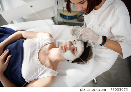 High angle view of beautician applying an alginic mask on a beautiful woman face at spa clinic 78035073