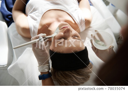 Overhead view of young woman relaxing on a massage table during beauty spa procedure and doctor cosmetologist applying alginate mask on her face 78035074