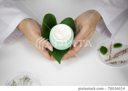 Hands hold jar with cream on green leaf, lab background with herbal ingredients 78036014