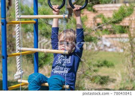 A blonde-haired child is playing on the playground, hanging on rings A blonde-haired child is playing on the playground, hanging on rings 78043841