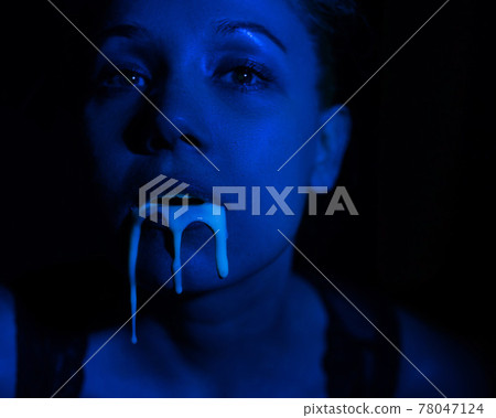A thick white liquid flows down the lips of a young European girl. Dripping down. Ultraviolet, blue light. Close-up portrait. Spit out.  78047124