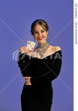 Brunette woman with bare shoulders, in black dress and shiny jewelry. Smiling, showing two aces, posing on purple background. Poker, casino. Close-up 78060139