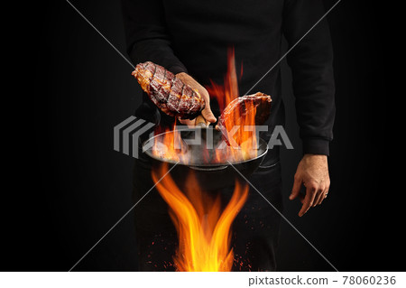 Young male dressed in jumper and jeans. He is holding burning wok pan with smoke above fire and frying two beef steaks, black background. Close up 78060236