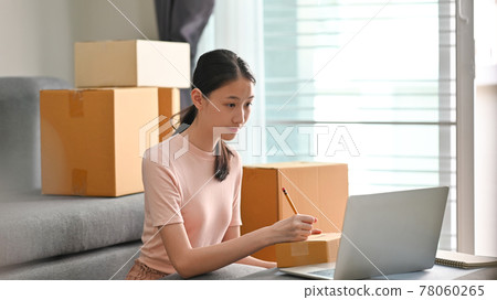 Beautiful girl writing costumer address on box while sitting at the modern living room. SME business startup. Beautiful girl writing costumer address on box while sitting at the modern living room. SME business startup. 78060265