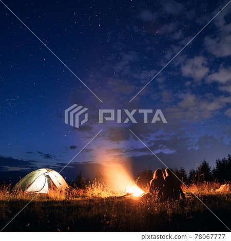 Young couple sitting near campfire under cloudy night sky. Young couple sitting near campfire under cloudy night sky. 78067777