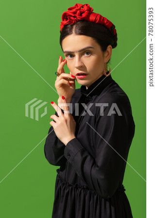 Close-up young beautiful woman in image of famous painter, artist Frida Kahlo on green background. Retro style, comparison of eras concept. Close-up young beautiful woman in image of famous painter, artist Frida Kahlo on green background. Retro style, comparison of eras concept. 78075393