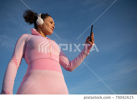Bottom view of awoman in pink sportswear with headphones and a mobile phone in her hand against the background of beautiful blue sky Bottom view of awoman in pink sportswear with headphones and a mobile phone in her hand against the background of beautiful blue sky 78082625