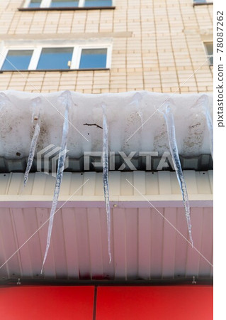 Long icicles hang dangerously down from the roof in close-up. Long icicles hang dangerously down from the roof in close-up. 78087262