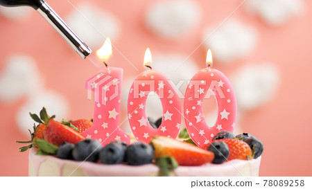 Birthday cake number 100, pink candle on beautiful cake with berries and lighter with fire against background of white clouds and pink sky. Close-up Birthday cake number 100, pink candle on beautiful cake with berries and lighter with fire against background of white clouds and pink sky. Close-up 78089258
