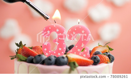 Birthday cake number 32, pink candle on beautiful cake with berries and lighter with fire against background of white clouds and pink sky. Close-up Birthday cake number 32, pink candle on beautiful cake with berries and lighter with fire against background of white clouds and pink sky. Close-up 78089261