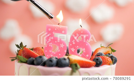 Birthday cake number 56, pink candle on beautiful cake with berries and lighter with fire against background of white clouds and pink sky. Close-up 78089291