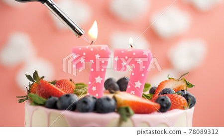 Birthday cake number 77, pink candle on beautiful cake with berries and lighter with fire against background of white clouds and pink sky. Close-up 78089312