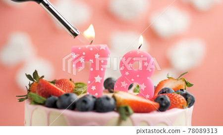 Birthday cake number 78, pink candle on beautiful cake with berries and lighter with fire against background of white clouds and pink sky. Close-up 78089313